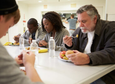 People Sitting At Table Eating Food In Homeless Shelter