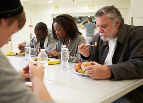 People Sitting At Table Eating Food In Homeless Shelter