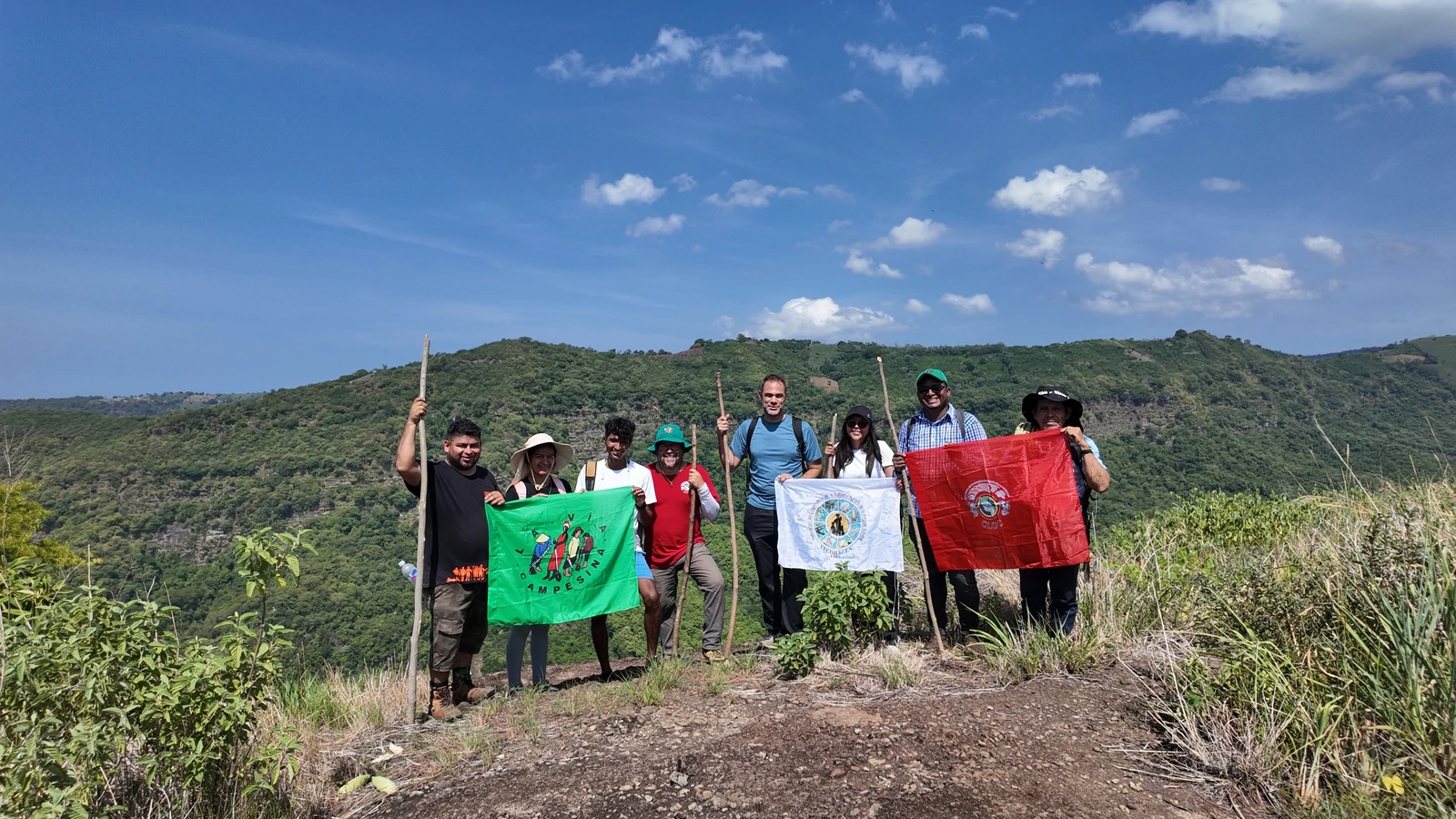 Members of a La Via Campesina solidarity delegation pose with organizational flags on a hilltop overlooking green valleys in El Salvador.