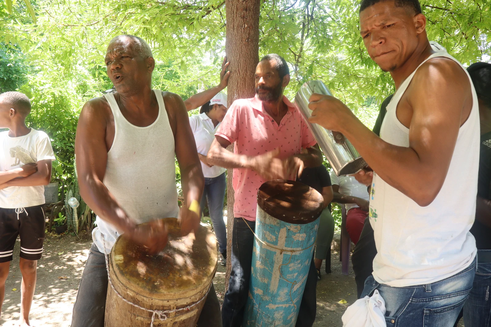 Three men playing traditional drums and percussion outdoors under trees