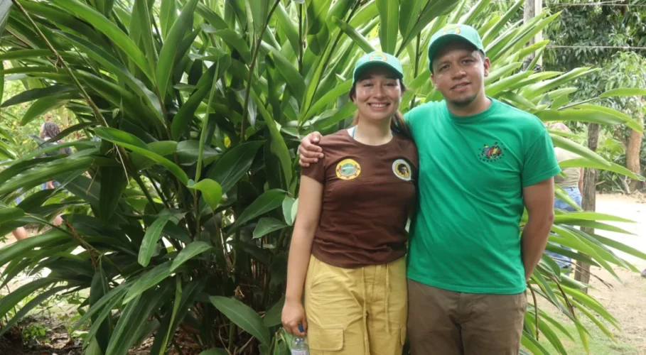 Two people smiling in front of tropical plants wearing La Via Campesina shirts