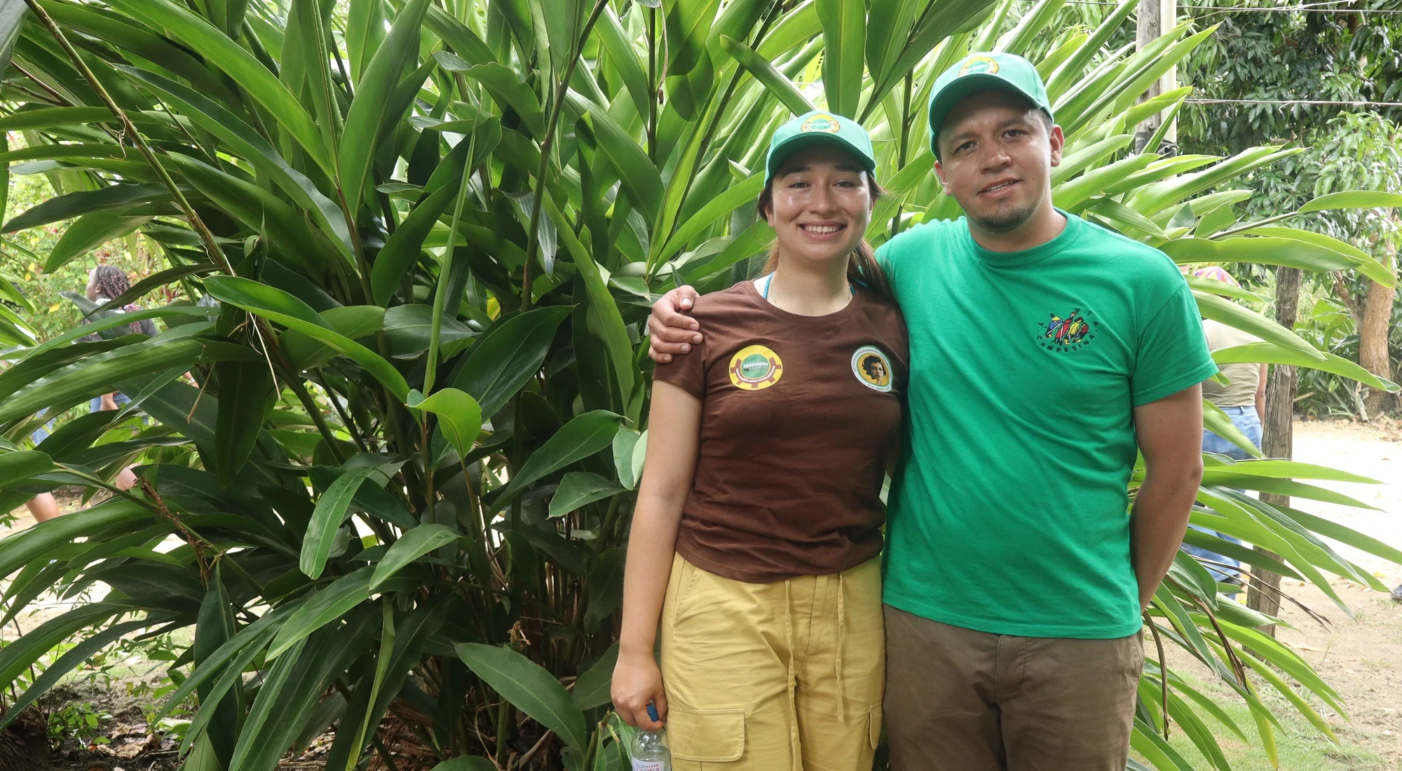 Two people smiling in front of tropical plants wearing La Via Campesina shirts