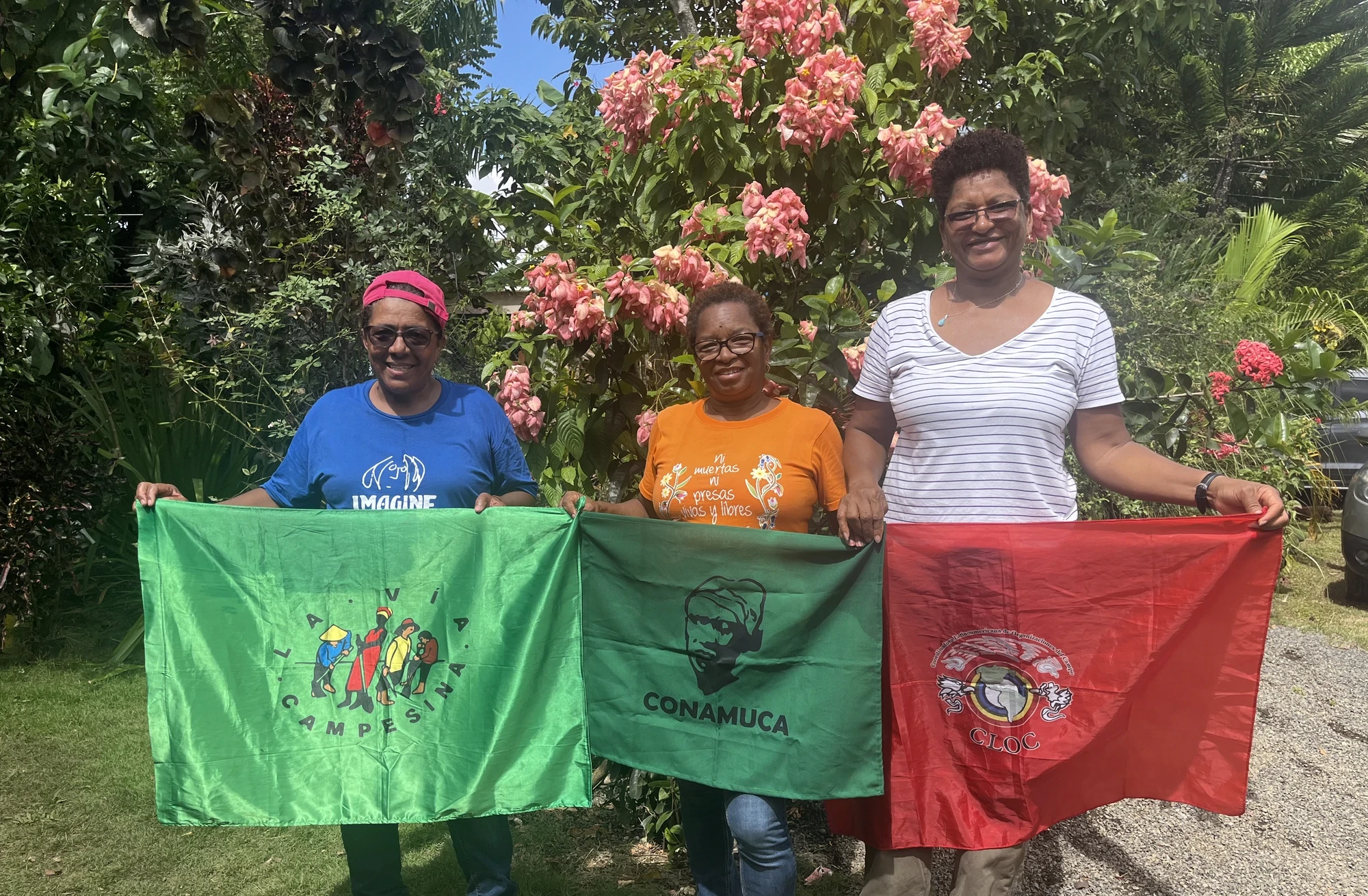 Three women holding La Via Campesina, CONAMUCA, and CLOC flags outdoors