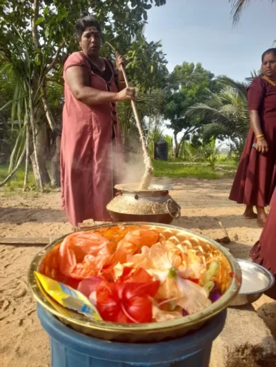 Woman stirring a large steaming pot outdoors with a bowl of hibiscus flowers nearby
