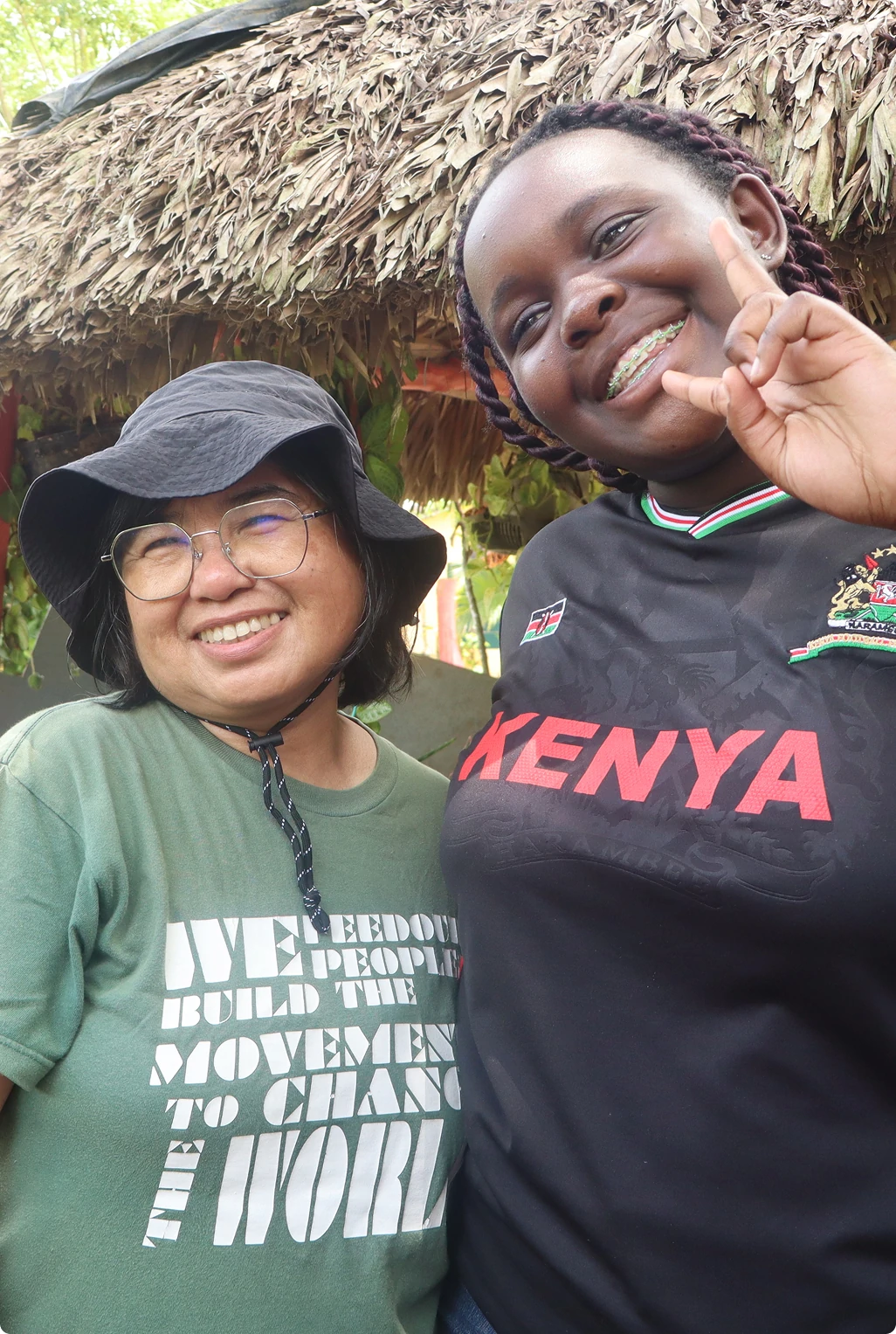 Two women smiling together outdoors under a thatched roof
